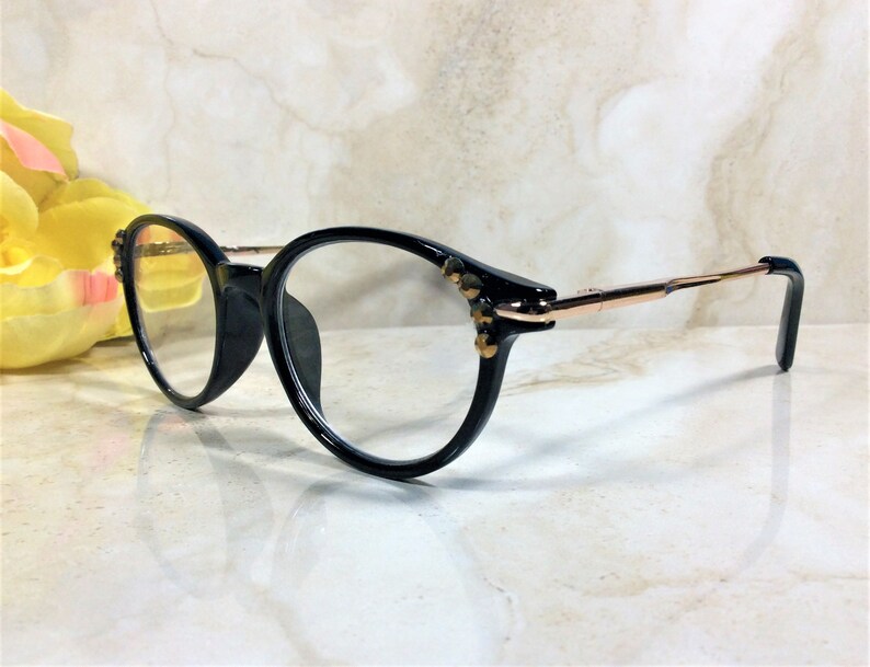 May include: Black eyeglasses with a round frame and gold-colored arms. The frames have decorative embellishments near the hinges. The lenses are clear. The glasses are set against a light-colored background.