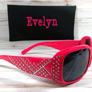 K&ouml;nnte beinhalten: Rosa Sonnenbrille mit Strasssteinen und schwarzen Gl&auml;sern. Die Sonnenbrille befindet sich in einem Etui mit dem Namen "Evelyn" darauf.