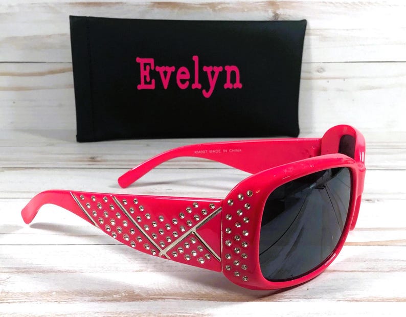 K&ouml;nnte beinhalten: Rosa Sonnenbrille mit Strasssteinen und schwarzen Gl&auml;sern. Die Sonnenbrille befindet sich in einem Etui mit dem Namen "Evelyn" darauf.