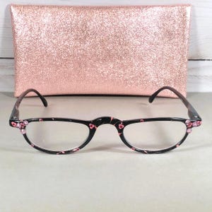Pu&ograve; includere: Occhiali neri con un delicato motivo floreale rosa sulla montatura e piccoli strass rosa agli angoli esterni. Gli occhiali sono posizionati davanti a una custodia rettangolare glitterata color oro rosa con una superficie ruvida e scintillante.
