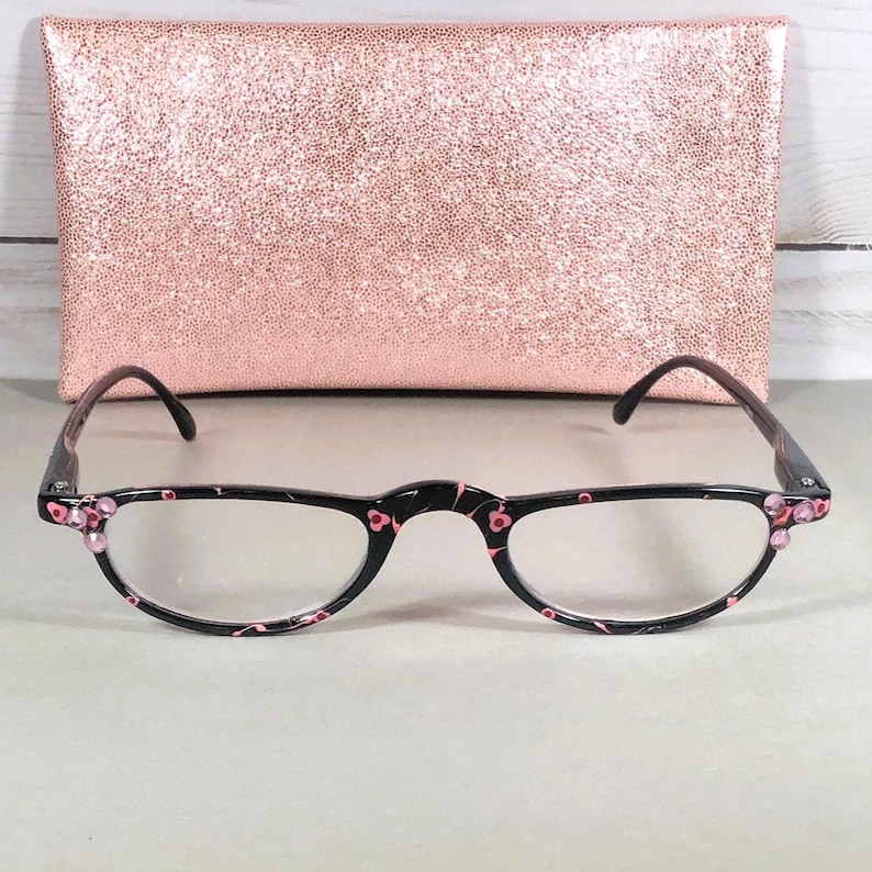 Pu&ograve; includere: Occhiali neri con un delicato motivo floreale rosa sulla montatura e piccoli strass rosa agli angoli esterni. Gli occhiali sono posizionati davanti a una custodia rettangolare glitterata color oro rosa con una superficie ruvida e scintillante.