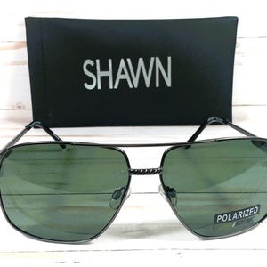 May include: Black aviator sunglasses with green lenses and a silver metal frame. The sunglasses have the word "POLARIZED" printed on the right lens. The sunglasses are sitting on a black case with the word "SHAWN" printed on it.