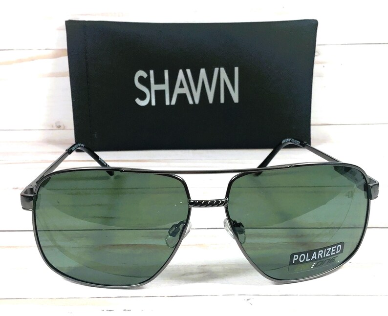 May include: Black aviator sunglasses with green lenses and a silver metal frame. The sunglasses have the word "POLARIZED" printed on the right lens. The sunglasses are sitting on a black case with the word "SHAWN" printed on it.