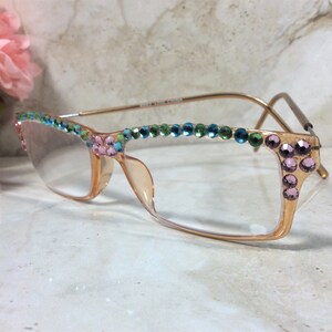 May include: A pair of eyeglasses with a light peach-colored frame and clear lenses. The top of the frame is adorned with a row of colorful rhinestones in shades of blue, green, and pink. The temples are gold-toned.