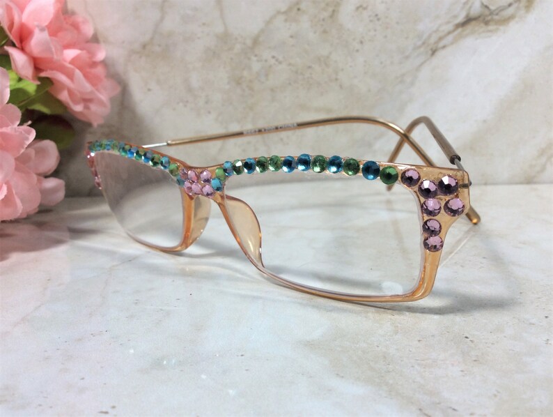 May include: A pair of eyeglasses with a light peach-colored frame and clear lenses. The top of the frame is adorned with a row of colorful rhinestones in shades of blue, green, and pink. The temples are gold-toned.