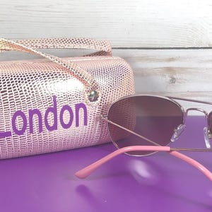 May include: Aviator sunglasses with pink arms and a matching pink case. The case, embossed with a reptile-like pattern, has the word "London" in purple lettering.