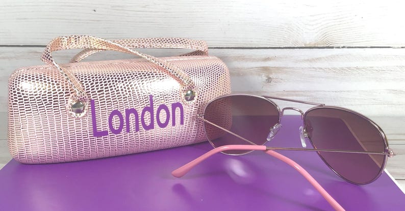 May include: Aviator sunglasses with pink arms and a matching pink case. The case, embossed with a reptile-like pattern, has the word "London" in purple lettering.
