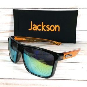 Op de afbeelding: Zwarte zonnebril met oranje monturen en blauwe gespiegelde glazen. De zonnebril ligt op een houten oppervlak naast een zwarte etui met het woord "Jackson" in oranje gedrukt.