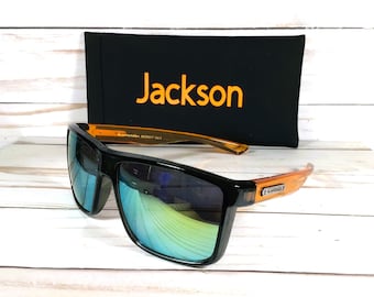May include: Black sunglasses with orange frames and blue mirrored lenses. The sunglasses are sitting on a wooden surface next to a black case with the word "Jackson" printed in orange.