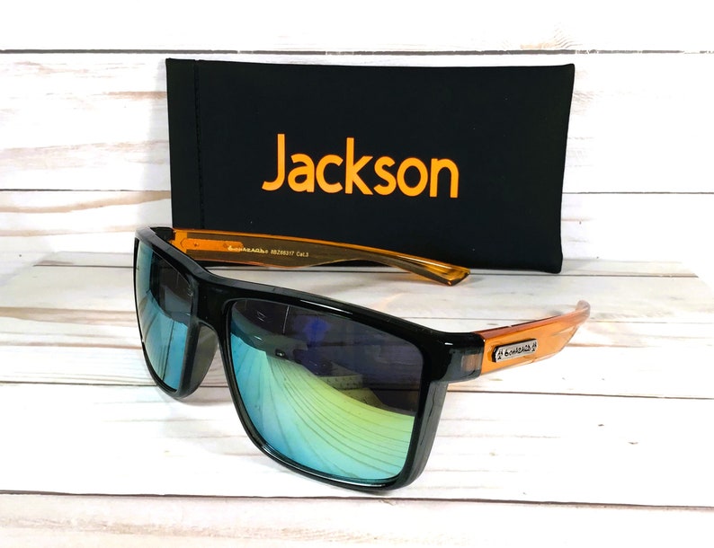 Op de afbeelding: Zwarte zonnebril met oranje monturen en blauwe gespiegelde glazen. De zonnebril ligt op een houten oppervlak naast een zwarte etui met het woord "Jackson" in oranje gedrukt.