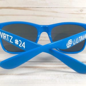 Puede incluir: Gafas de sol de color azul brillante con lentes oscuras. La patilla izquierda presenta el texto blanco WIRTZ #24, mientras que la patilla derecha muestra el texto blanco ULTIMATE junto a un icono gr&aacute;fico blanco de un bal&oacute;n de voleibol.