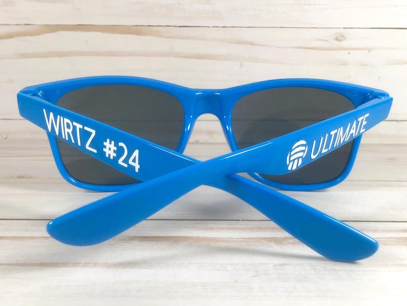 Puede incluir: Gafas de sol de color azul brillante con lentes oscuras. La patilla izquierda presenta el texto blanco WIRTZ #24, mientras que la patilla derecha muestra el texto blanco ULTIMATE junto a un icono gr&aacute;fico blanco de un bal&oacute;n de voleibol.