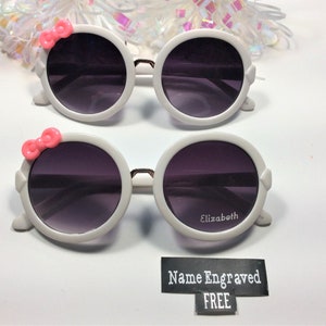 May include: White round sunglasses with pink bows on the sides. The sunglasses have dark lenses and the words "Elizabeth" and "Name Engraved FREE" are printed on the bottom.