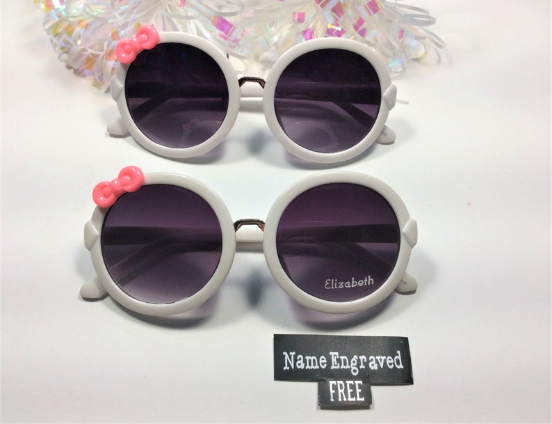 May include: White round sunglasses with pink bows on the sides. The sunglasses have dark lenses and the words "Elizabeth" and "Name Engraved FREE" are printed on the bottom.