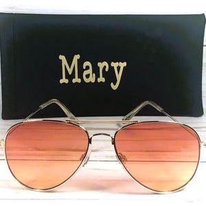 May include: Gold-framed aviator sunglasses with orange gradient lenses, in front of a black case. The case is personalised with the name "Mary" in gold lettering.