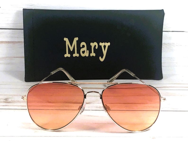 May include: Gold-framed aviator sunglasses with orange gradient lenses, in front of a black case. The case is personalised with the name "Mary" in gold lettering.
