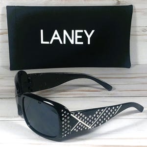 May include: Black sunglasses with a decorative pattern of rhinestones and a white X design on the temples. The sunglasses are paired with a black case that has the name "LANEY" printed in white.