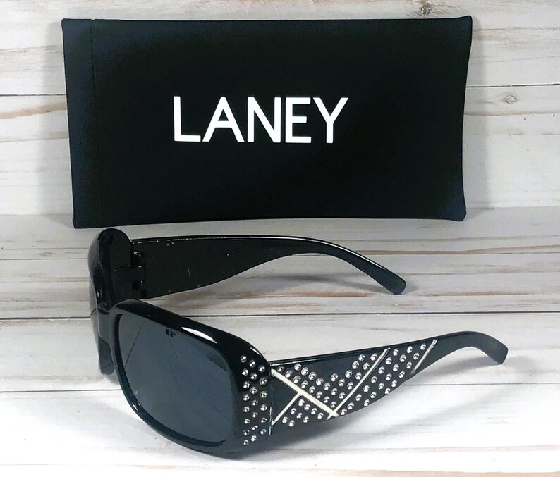 May include: Black sunglasses with a decorative pattern of rhinestones and a white X design on the temples. The sunglasses are paired with a black case that has the name "LANEY" printed in white.