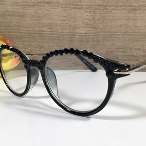 Puede incluir: Gafas negras con una fila decorativa de peque&ntilde;as cuentas negras facetadas a lo largo del borde superior de la montura. Las gafas tienen lentes transparentes y patillas finas de metal plateado que se extienden desde los lados de la montura.
