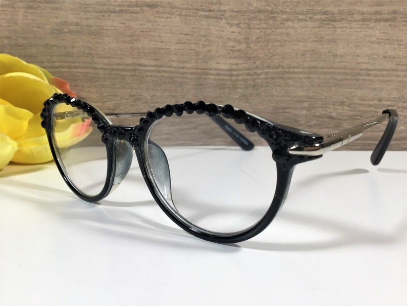 Puede incluir: Gafas negras con una fila decorativa de peque&ntilde;as cuentas negras facetadas a lo largo del borde superior de la montura. Las gafas tienen lentes transparentes y patillas finas de metal plateado que se extienden desde los lados de la montura.