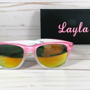 Puede incluir: Gafas de sol rosa y blancas con lentes de color naranja degradado. Las gafas de sol est&aacute;n sobre una superficie de madera blanca. El nombre "Layla" est&aacute; escrito en rosa sobre un fondo negro.