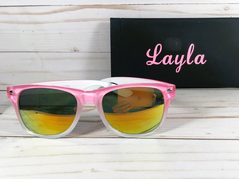 Puede incluir: Gafas de sol rosa y blancas con lentes de color naranja degradado. Las gafas de sol est&aacute;n sobre una superficie de madera blanca. El nombre "Layla" est&aacute; escrito en rosa sobre un fondo negro.