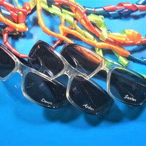 May include: Three pairs of black sunglasses with clear frames. The sunglasses are on a blue background with colourful curly ribbons. The sunglasses are personalised with the names "Jaxon", "Ayden", and "Devyn".