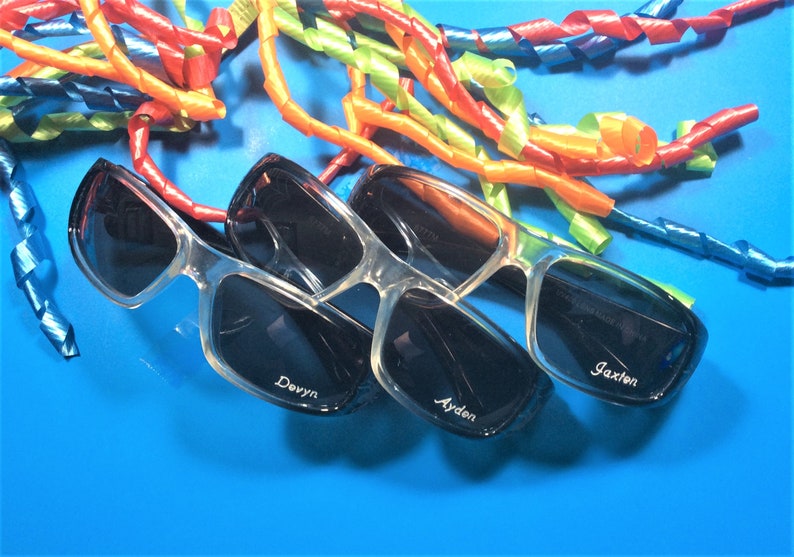 May include: Three pairs of black sunglasses with clear frames. The sunglasses are on a blue background with colourful curly ribbons. The sunglasses are personalised with the names "Jaxon", "Ayden", and "Devyn".