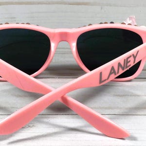 Op de afbeelding: Roze zonnebril met zwarte glazen en de naam "LANEY" gedrukt op de zijkant.