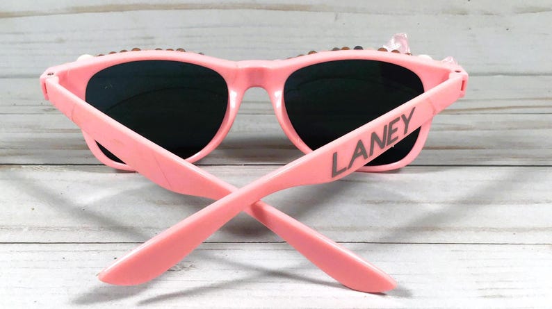 Op de afbeelding: Roze zonnebril met zwarte glazen en de naam "LANEY" gedrukt op de zijkant.