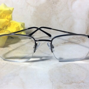 May include: Semi-rimless spectacles with black frames and decorative black detailing on the corners. The lenses are clear, and the arms are thin. The glasses are set against a soft, marble-like background with a yellow flower.