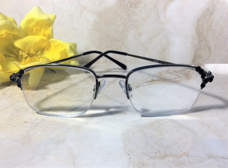 May include: Semi-rimless spectacles with black frames and decorative black detailing on the corners. The lenses are clear, and the arms are thin. The glasses are set against a soft, marble-like background with a yellow flower.