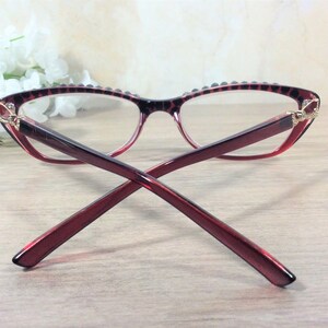 May include: Cat-eye style spectacles in red and black, featuring a decorative pattern on the frame. The lenses are clear, and the frame has a gradient effect. The arms of the glasses are a solid red colour.