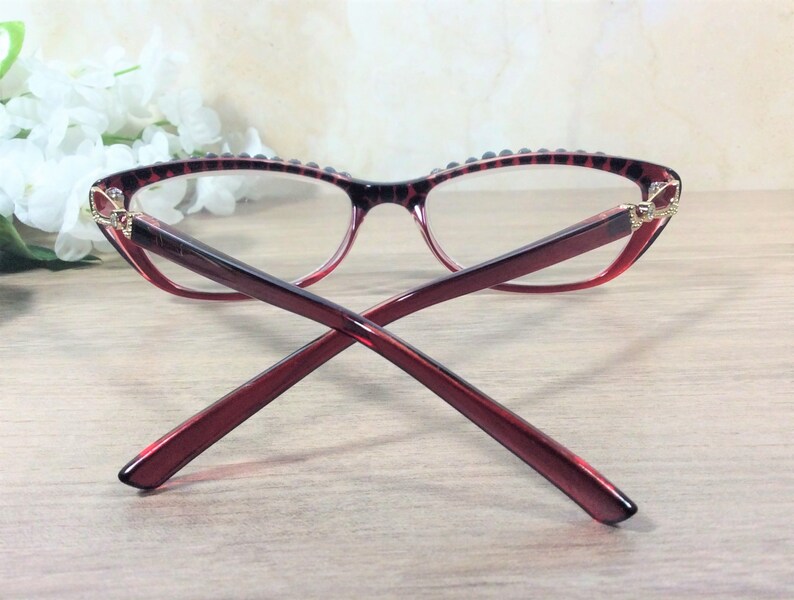 May include: Cat-eye style spectacles in red and black, featuring a decorative pattern on the frame. The lenses are clear, and the frame has a gradient effect. The arms of the glasses are a solid red colour.