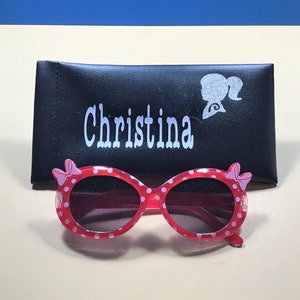 May include: Pink and white polka dot sunglasses with a black case. The case has the name "Christina" and a silhouette of a girl with a ponytail on it.