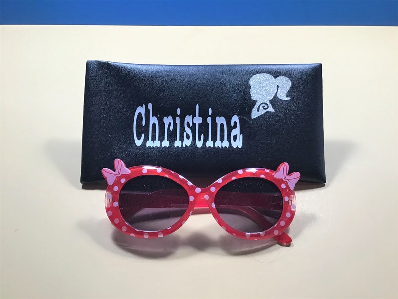 May include: Pink and white polka dot sunglasses with a black case. The case has the name "Christina" and a silhouette of a girl with a ponytail on it.