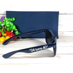 K&ouml;nnte beinhalten: Marineblaue Sonnenbrille mit dem wei&szlig;en Text "04 turns 40". Eine marineblaue H&uuml;lle und bunte B&auml;nder befinden sich im Hintergrund. Die Sonnenbrille hat einen klassischen Stil.