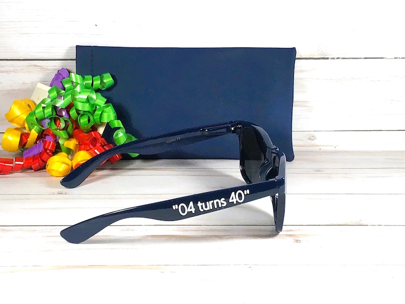 K&ouml;nnte beinhalten: Marineblaue Sonnenbrille mit dem wei&szlig;en Text "04 turns 40". Eine marineblaue H&uuml;lle und bunte B&auml;nder befinden sich im Hintergrund. Die Sonnenbrille hat einen klassischen Stil.