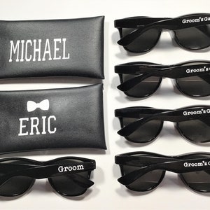 May include: Five pairs of black sunglasses with white text. The sunglasses have a classic aviator style. The text on the sunglasses reads "Groom's Gang" and "Groom". Two black leather cases with white text are also included. The text on the cases reads "Michael" and "Eric" with a bowtie symbol.