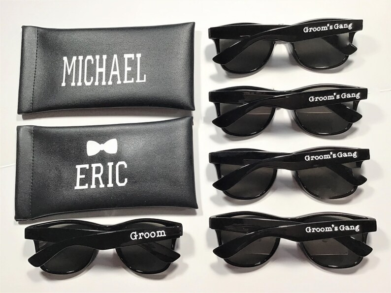 May include: Five pairs of black sunglasses with white text. The sunglasses have a classic aviator style. The text on the sunglasses reads "Groom's Gang" and "Groom". Two black leather cases with white text are also included. The text on the cases reads "Michael" and "Eric" with a bowtie symbol.