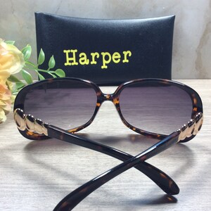 Puede incluir: Gafas de sol de carey con detalles dorados en las patillas. Las gafas tienen lentes grises oscuras y se muestran con una funda negra que dice "Harper" en letras amarillas.