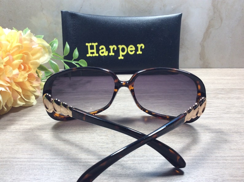 Puede incluir: Gafas de sol de carey con detalles dorados en las patillas. Las gafas tienen lentes grises oscuras y se muestran con una funda negra que dice "Harper" en letras amarillas.