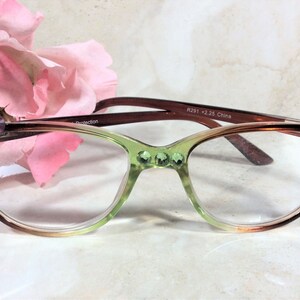 May include: Cat-eye reading glasses with a gradient green and brown frame, adorned with pink rhinestones. The glasses have clear lenses and are set against a soft pink flower background.
