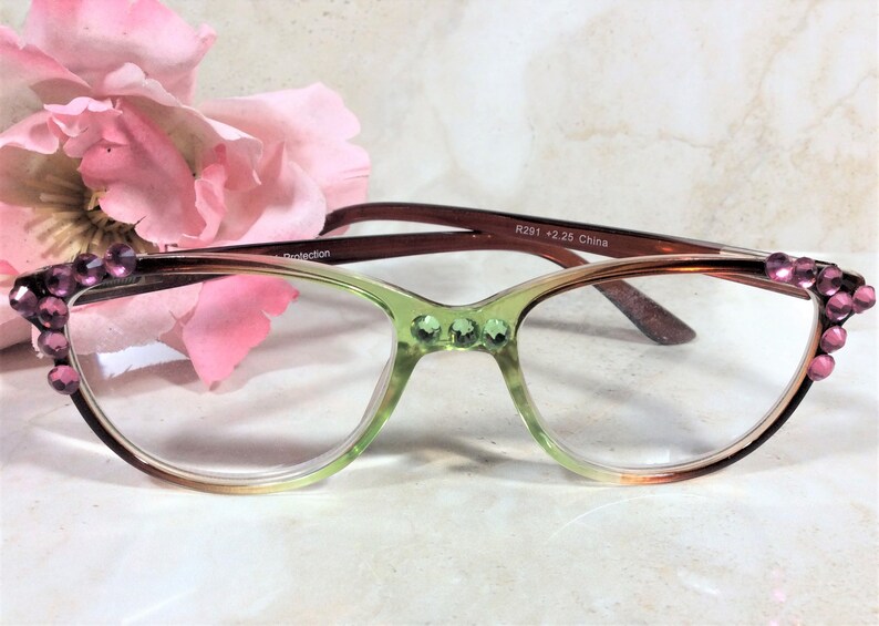 May include: Cat-eye reading glasses with a gradient green and brown frame, adorned with pink rhinestones. The glasses have clear lenses and are set against a soft pink flower background.