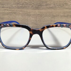 May include: A pair of eyeglasses featuring a tortoiseshell pattern frame, blue arms, and clear lenses. The frame corners are decorated with pink and blue rhinestones. The glasses are set against a white surface.
