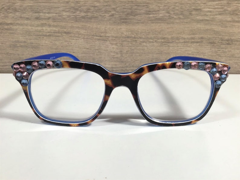 May include: A pair of eyeglasses featuring a tortoiseshell pattern frame, blue arms, and clear lenses. The frame corners are decorated with pink and blue rhinestones. The glasses are set against a white surface.