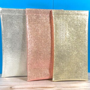 May include: Three rectangular pouches with a glittery, textured finish. The left pouch is silver, the middle pouch is rose gold, and the right pouch is gold. Each pouch has a smooth, flat top edge and a vertical orientation with a shimmering surface.