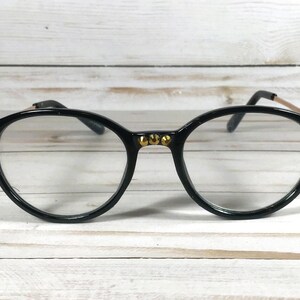 May include: Black round-frame glasses featuring gold-tone metal accents on the bridge and outer corners. The frames have clear lenses and thin gold-tone metal temples. The bridge and corners are decorated with small, circular gold-tone studs.