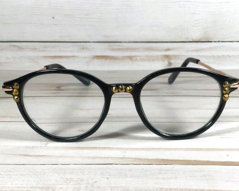 May include: Black round-frame glasses featuring gold-tone metal accents on the bridge and outer corners. The frames have clear lenses and thin gold-tone metal temples. The bridge and corners are decorated with small, circular gold-tone studs.