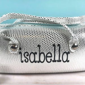 May include: Silver cosmetic bag with a textured, dotted pattern and a matching handle. The name "Isabella" is embroidered in black script on the front. The bag has silver hardware.